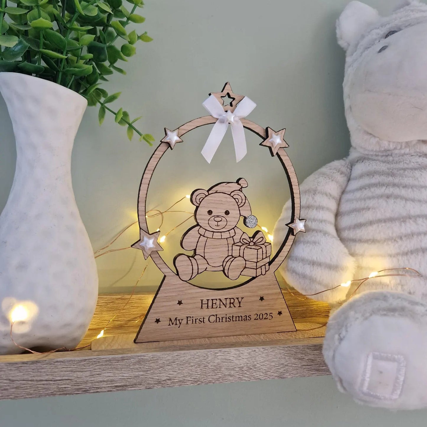 Wooden plaque with a teddy bear design and 'My First Christmas' text, surrounded by decorative items on a shelf.