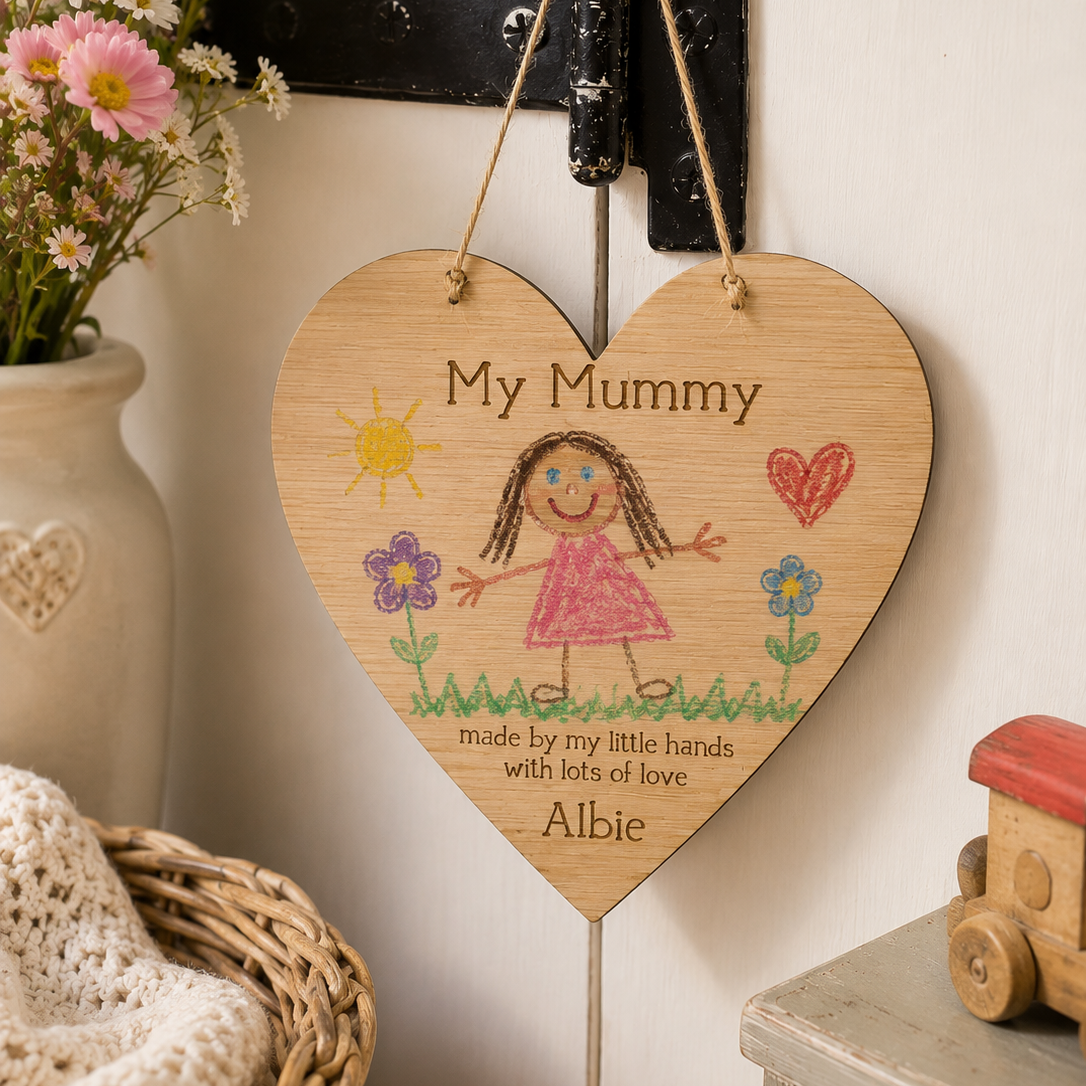 A heart-shaped wooden hanging plaque engraved with the message 'My Mummy made by my little hands with lots of love. Albie' and decorated with a childlike drawing of a girl, flowers, and a sun.