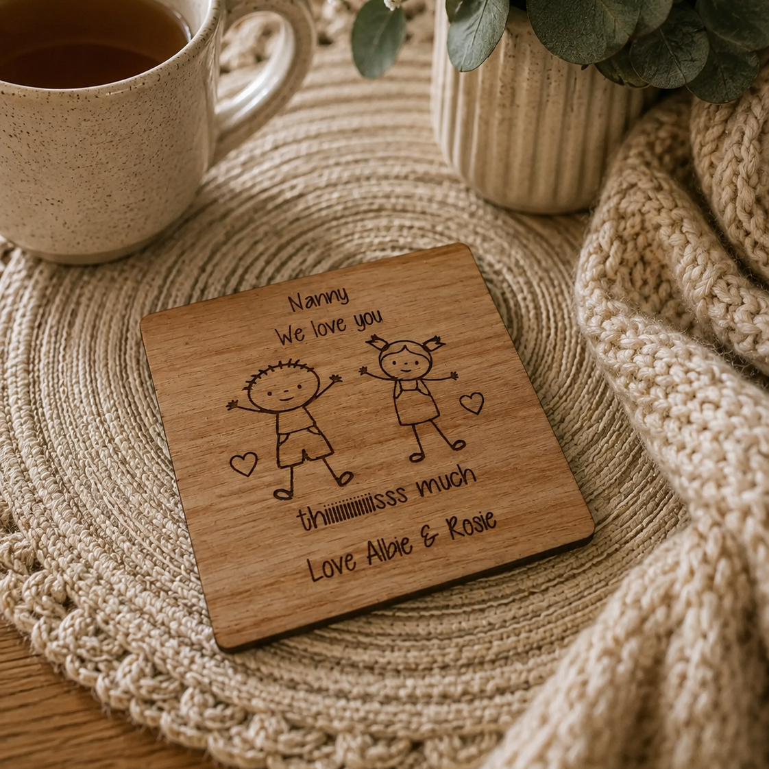 A square wooden coaster with engraved cartoon children and the message 'Nanny We love you thummmss much Love Alice & Rose' placed on a woven placemat beside a mug of tea, flowers, and a knitted blanket.