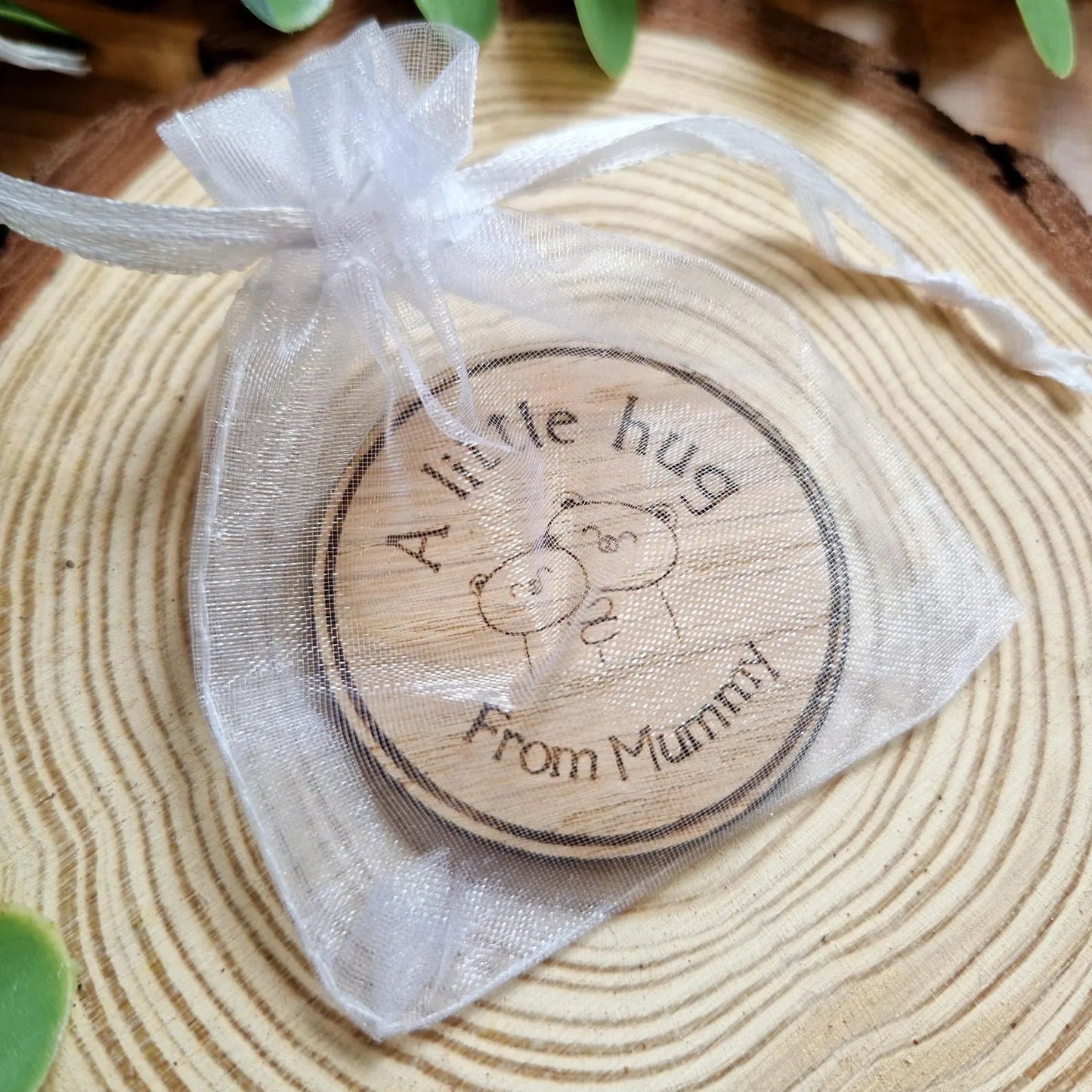 Wooden round token with 'A little hug from Mummy' text on a white ribbon, placed on a wooden surface with green leaves.