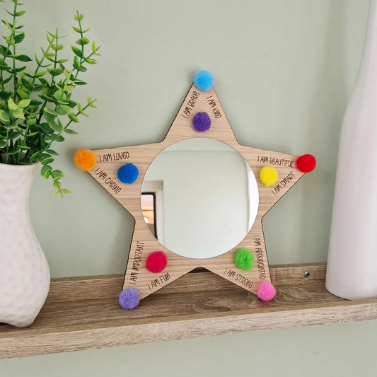 Star-shaped mirror with colorful pom-poms on a wooden shelf against a light green wall.