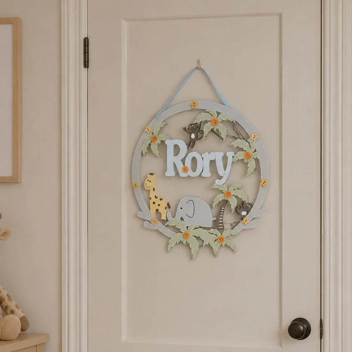 A personalized wooden nursery door hanger featuring the name 'Rory' in white letters, decorated with safari animal illustrations including a giraffe, elephant, and zebra, hanging on a cream-colored nursery door.