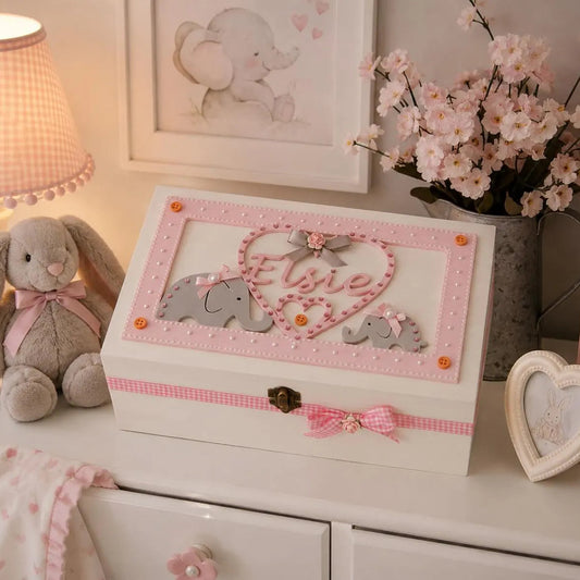 A decorative white wooden keepsake box with pink gingham trim, a heart-shaped cutout, and a pink bow, styled on a white dresser beside a plush bunny, lamp, books, and flowers.